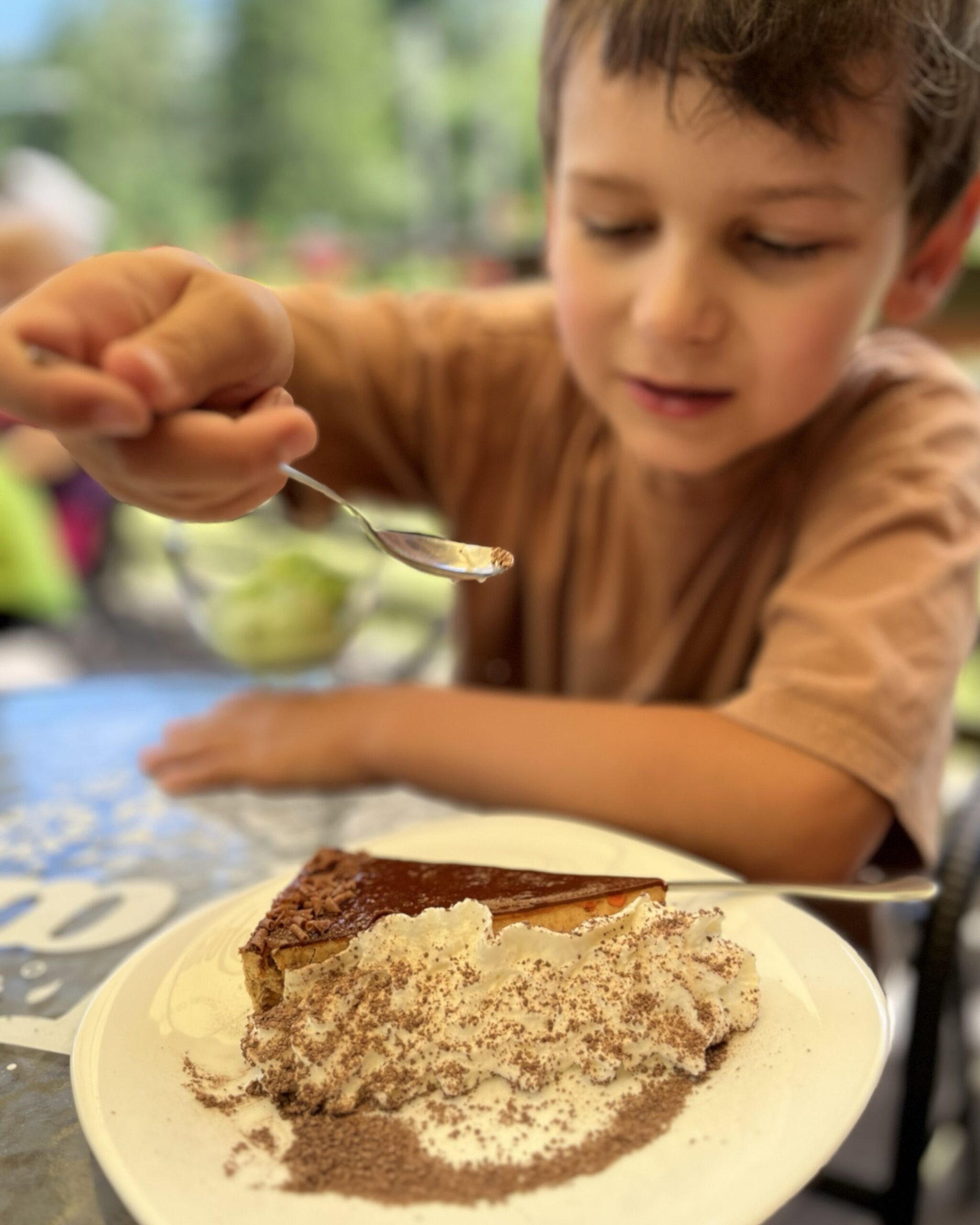 Chłopiec je sernik z bitą śmietaną i kakao w kawiarni z widokiem, obok puchar lodów.